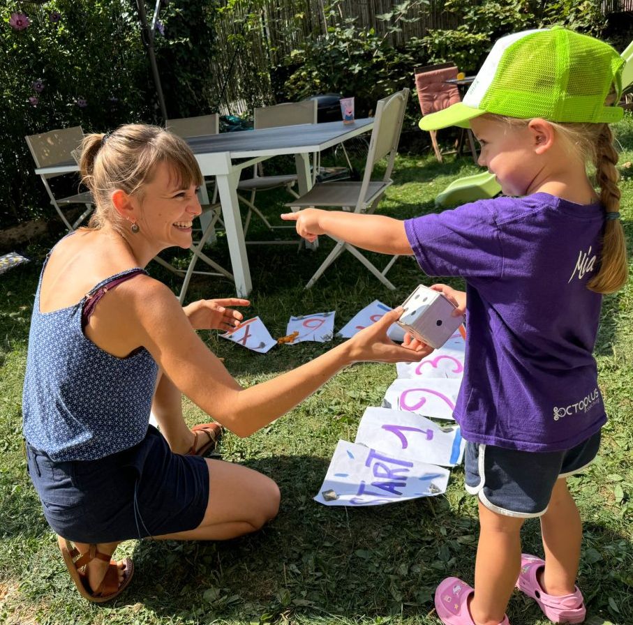 Enfant apprenant l'anglais en s'amusant dans un cadre naturel chez Smile & Move Une éducatrice de Smile & Move échange avec une petite fille souriante lors d'une activité ludique en plein air. L'enfant, portant une casquette verte, participe à un jeu éducatif pour découvrir l'anglais dans un environnement naturel et bienveillant.